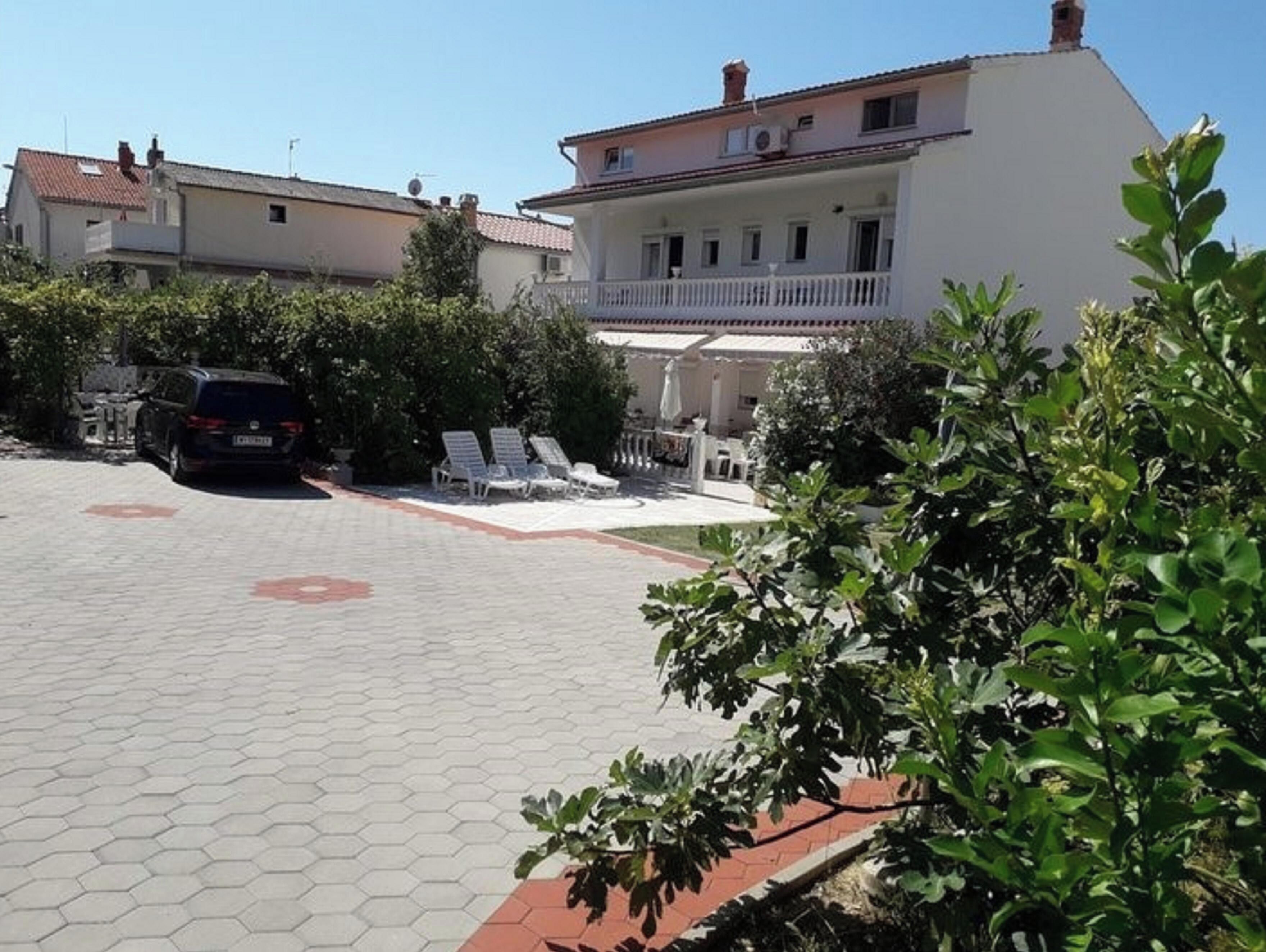 A residential building with a paved driveway, a car parked in the driveway, and a balcony with outdoor furniture.
