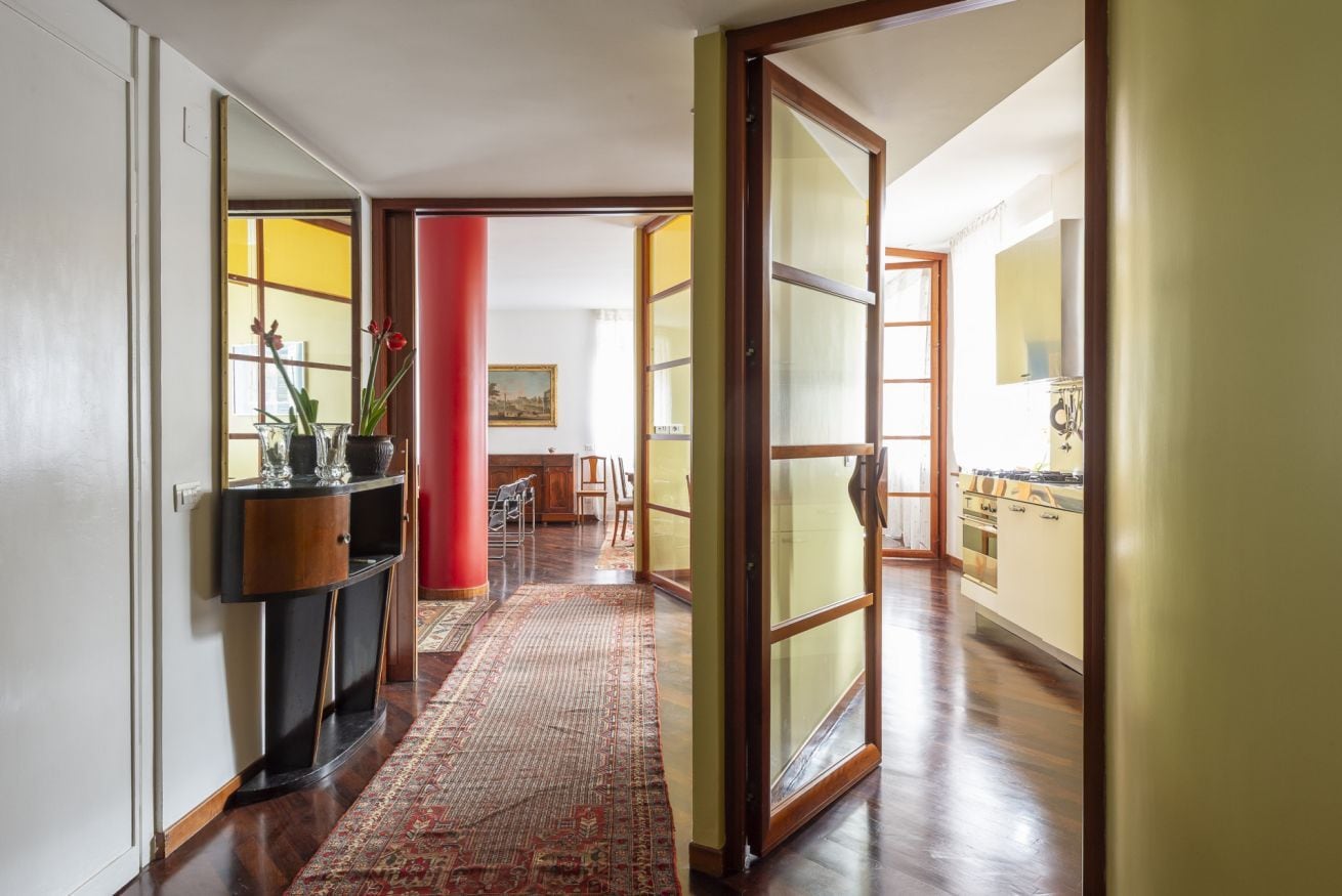 A hallway with a wooden floor, a red pillar, and a glass door leading to a kitchen area.