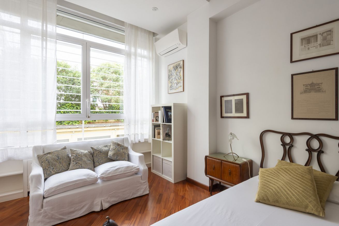 A bedroom with a bed, a white sofa, a wooden nightstand, a bookshelf, and a window with curtains.