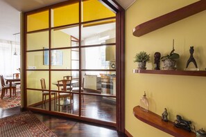 Habitació amb porta corredissa de vidre que dona a una cuina, zona de menjador amb caires i una estant amb objectes decoratius.