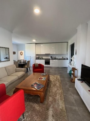 A modern living room with a sofa, red armchair, and coffee table, leading to a kitchen with white cabinets and a dining area.