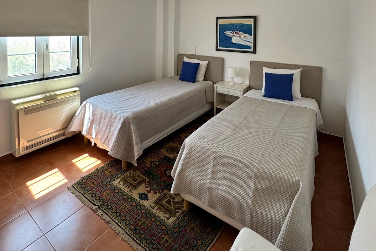 Une chambre d’hôtel avec deux lits, une fenêtre avec des stores, un radiateur, un tapis et un tableau au mur.