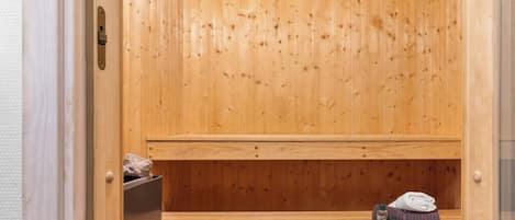 Una sauna de madera con un banco, un estante con piedras, y una toalla y una botella en el suelo.