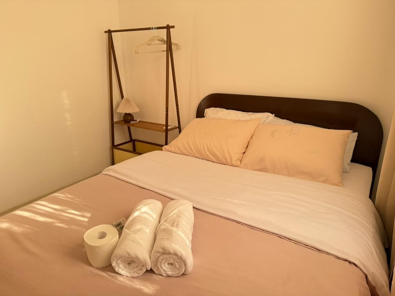 Una cama con cabecera, una mesita de noche con lámpara y toallas enrolladas sobre la cama.