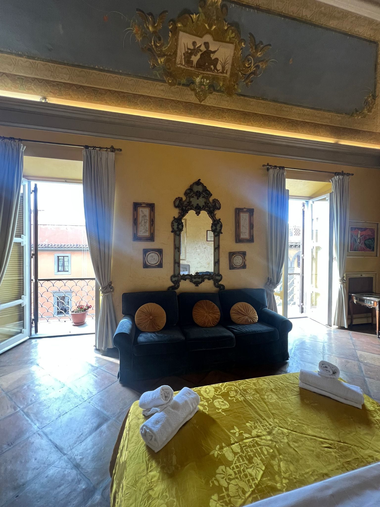 Una habitación de hotel con una cama grande, un sofá oscuro, un espejo decorativo y un balcón con vistas.