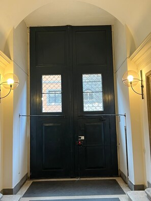 Uma porta dupla preta com uma pequena janela em cada porta, um puxador e uma maçaneta vermelha. Existem dois apliques de parede em ambos os lados da porta.