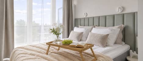 Un dormitorio con cama, mesitas de noche, una ventana con cortinas, y una mesita con una bandeja que sostiene un libro y un florero con flores.