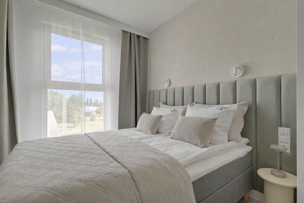 Makuuhuoneessa on suuri sänky, yöpöytä, seinään kiinnitetty valaisin ja ikkuna, jossa on verhot.