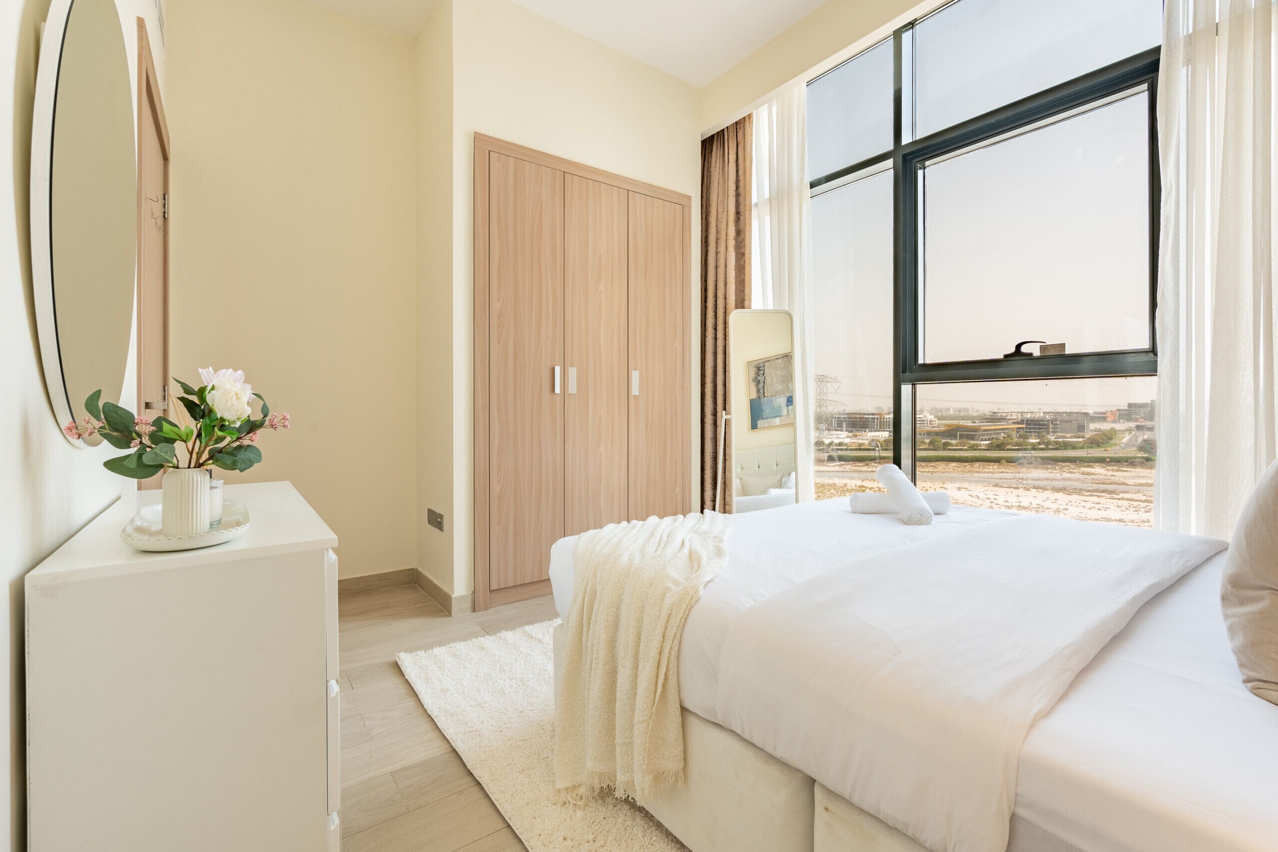 A bedroom with a large window, a bed, a bedside table with a mirror, and a wardrobe.