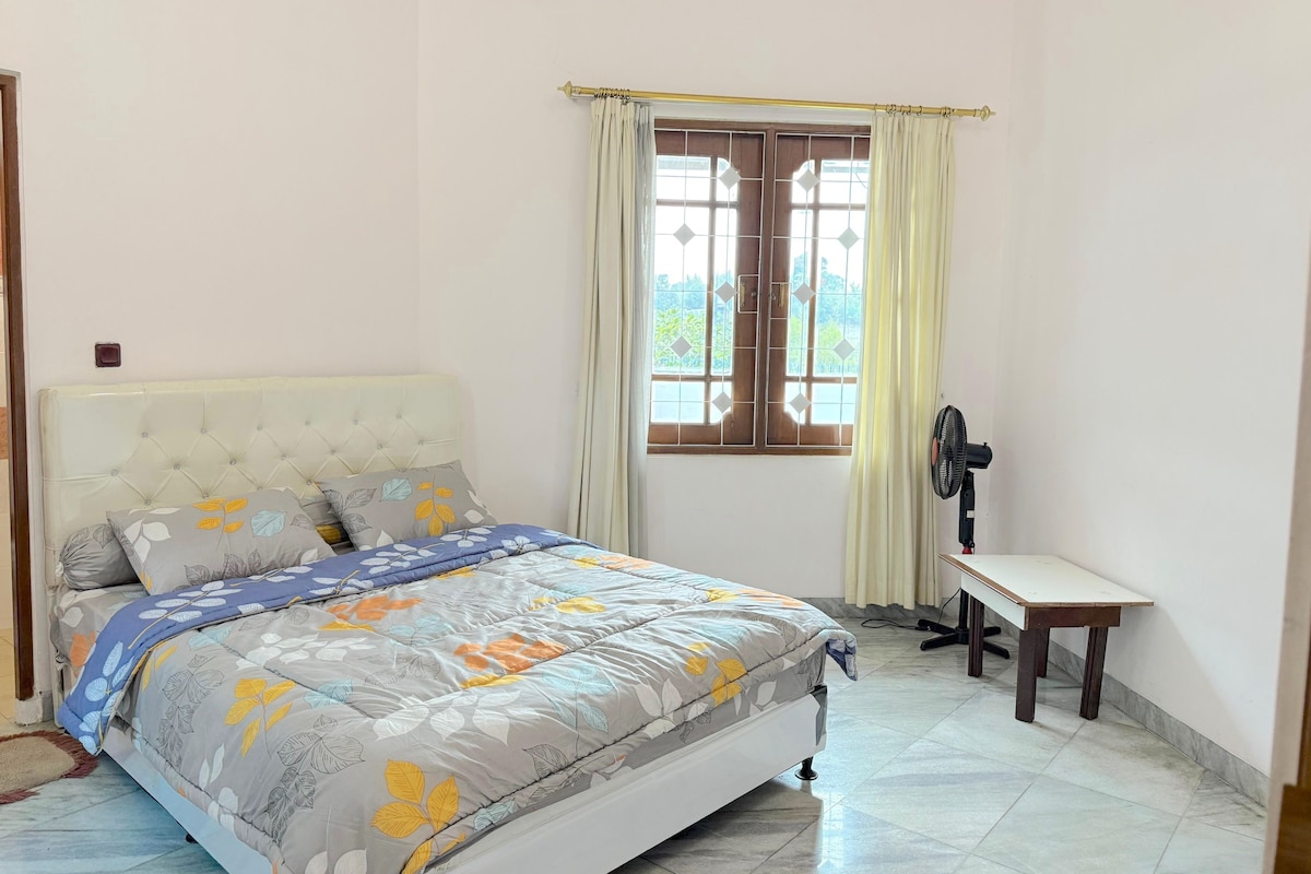 A bedroom with a bed, a window with curtains, a desk, and a fan.