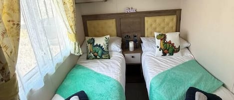 Habitación compacta con dos camas, cada una con colcha verde, una almohada, cabecera, mesita de noche y un estante pequeño sobre las camas.