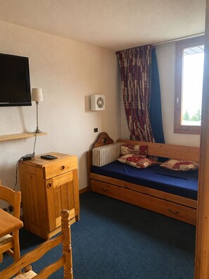 Ein kleines Zimmer mit einem Bett, einem Schreibtisch, einem Stuhl, einem Fernseher und einem fenster mit Vorhängen.