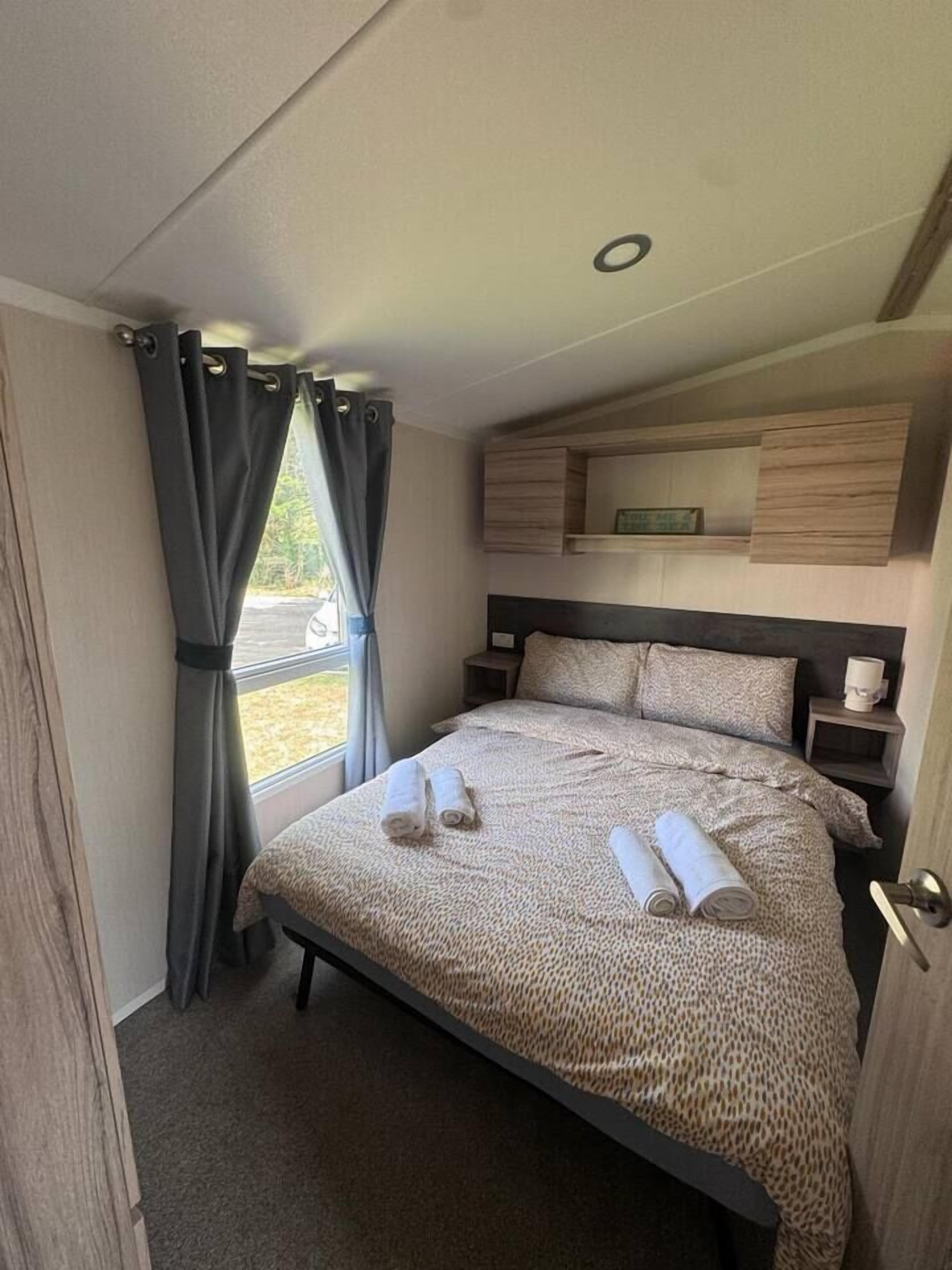 A bedroom with a bed, curtains, a headboard, a bedside table, and a wall-mounted shelf.