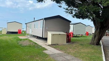 Uma casa móvel com um pequeno depósito, um caminho de entrada pavimentado e uma árvore grande.