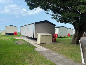 A mobile home with a small attached utility shed, a paved driveway, and a large tree.