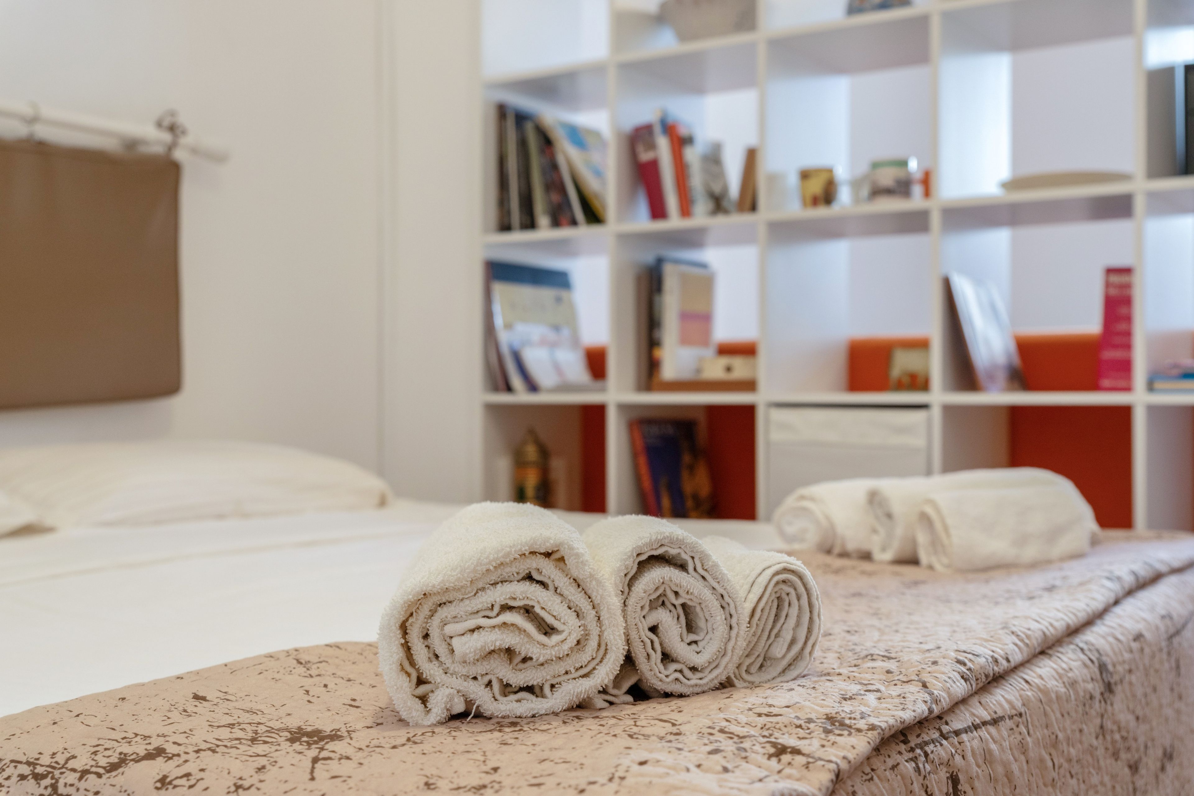 A neatly made bed with rolled white towels and a bookshelf in the background.
