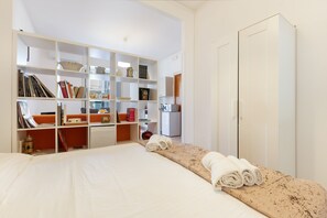 Une chambre d’hôtel comprenant un lit, une grande armoire blanche, un bureau avec une chaise et une bibliothèque remplie de livres et d’objets décoratifs.