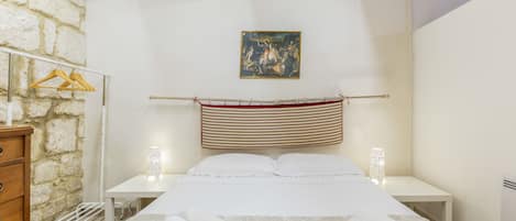 A bedroom with a bed, two bedside tables, a wooden dresser, a stone wall, a painting, and a towel rack.