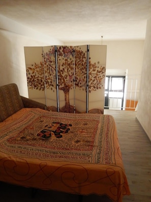 Un dormitori amb un llit cobert per un llençol estampat, un mural decoratiu amb un arbre a la paret, i vistes al balcó exterior.