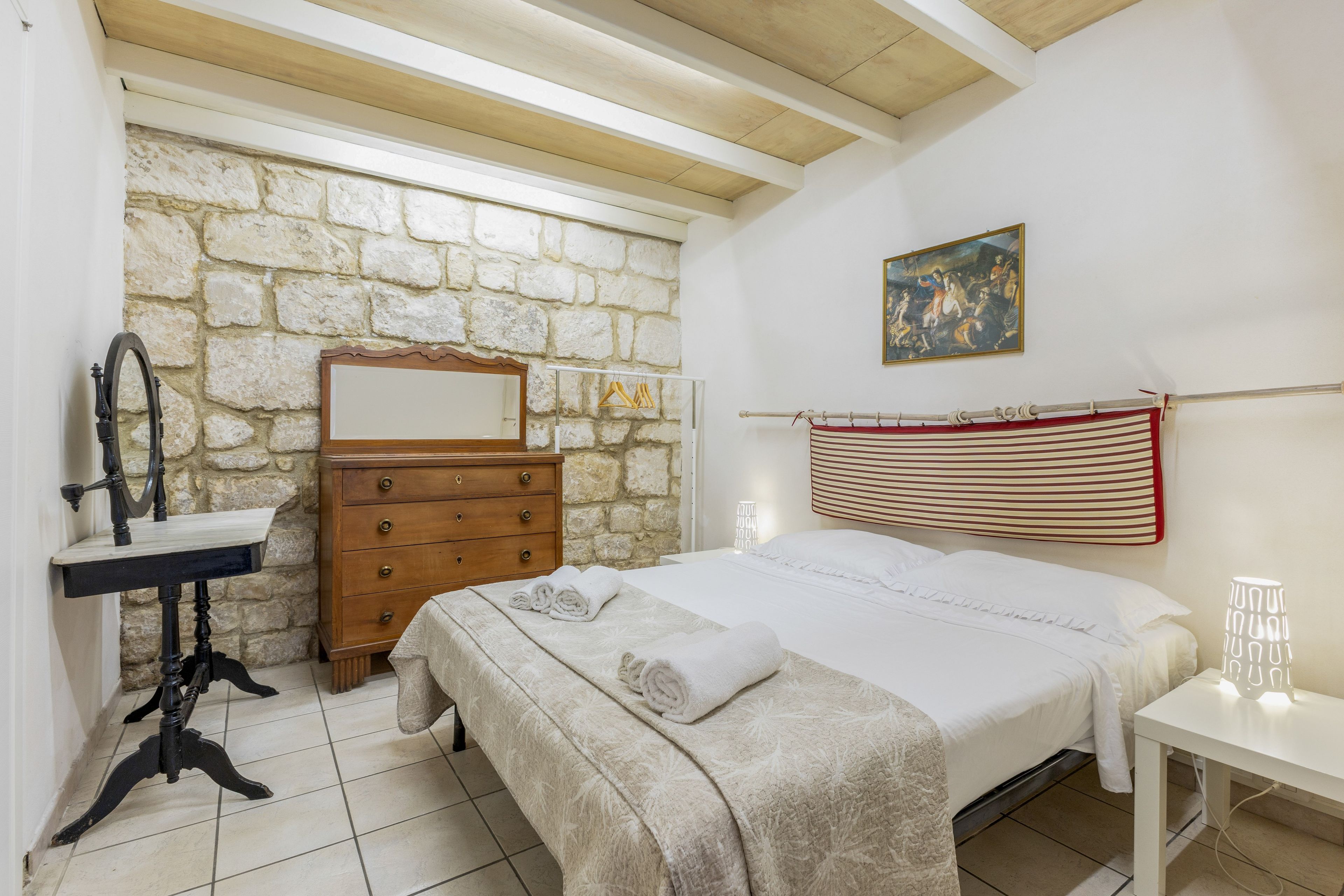 A bedroom with a stone wall, wooden ceiling beams, a bed with white bedding, a dresser, a mirror, and a painting.