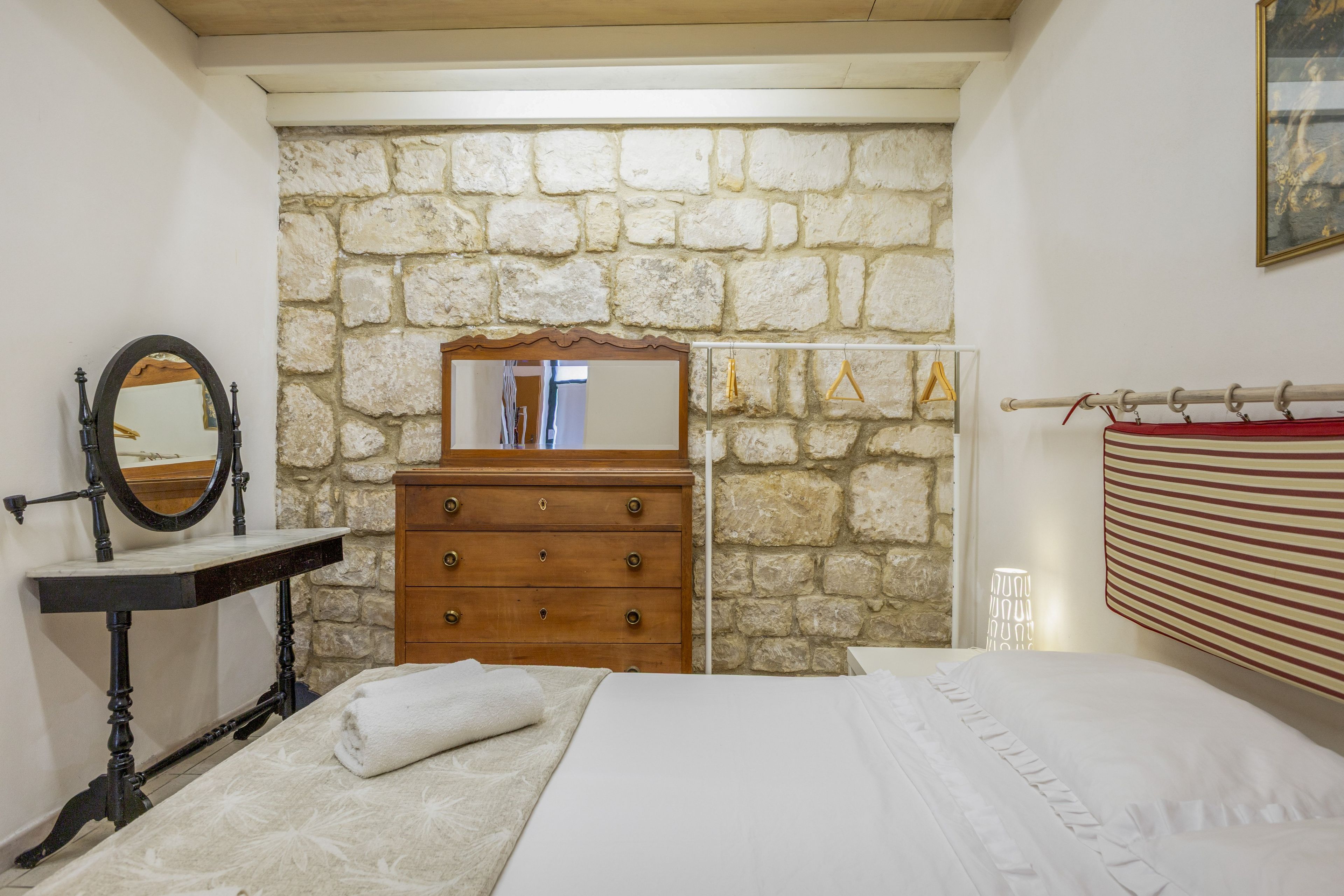 A bedroom with a stone wall, a wooden dresser, a mirror, a bed, and a hanging light fixture.