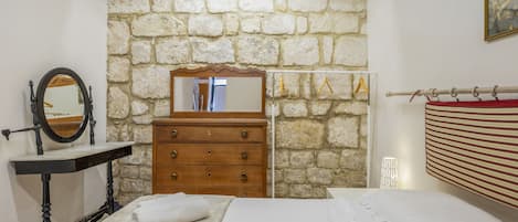 A bedroom with a stone wall, a wooden dresser, a mirror, a bed, and a hanging light fixture.