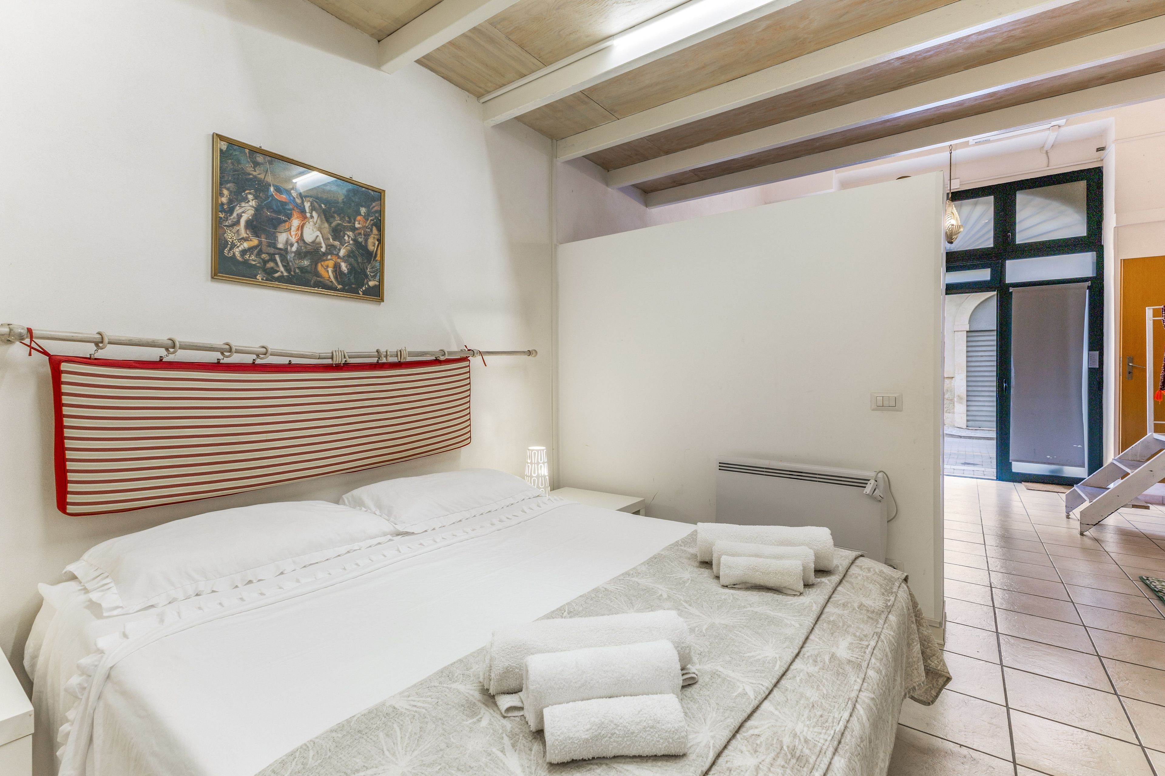 A hotel room with a bed, a painting on the wall, and a view of the outside.