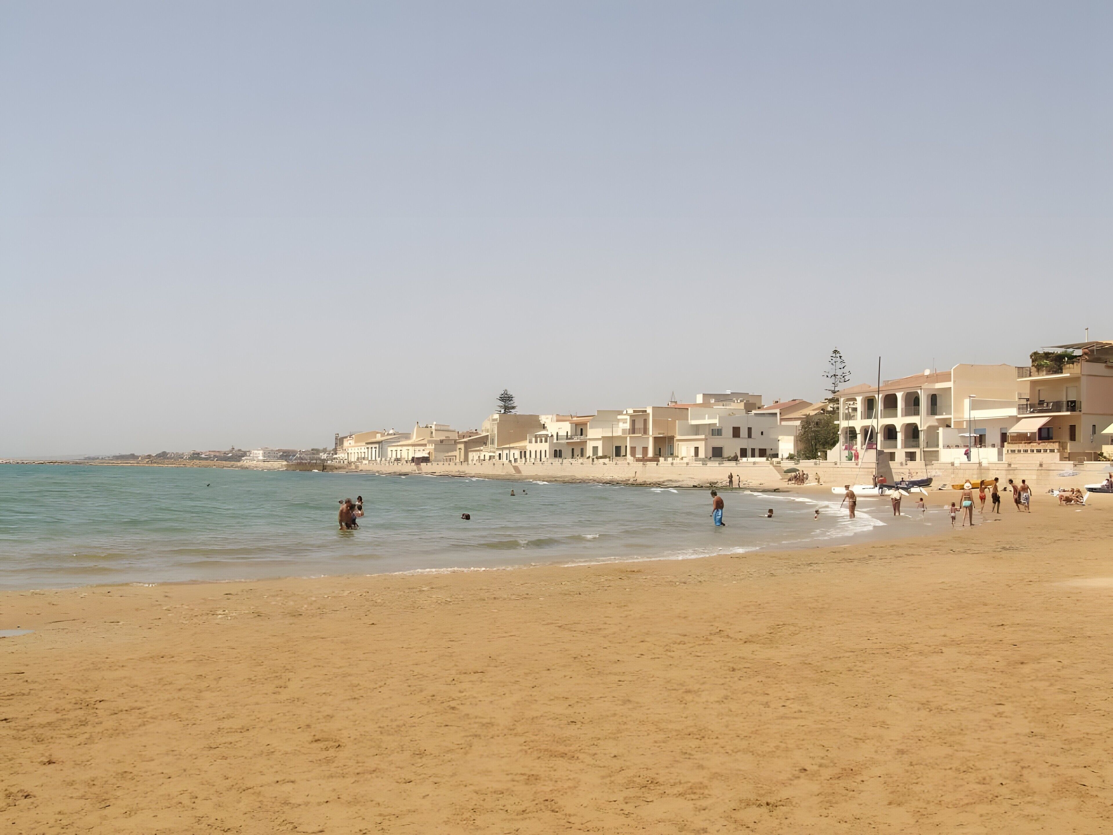 Una spiaggia con edifici sullo sfondo, persone sulla sabbia e acqua calma.