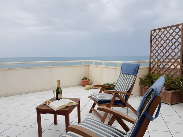 A terrace with two striped lounge chairs, a table with a book and a bottle, and a view of the sea.