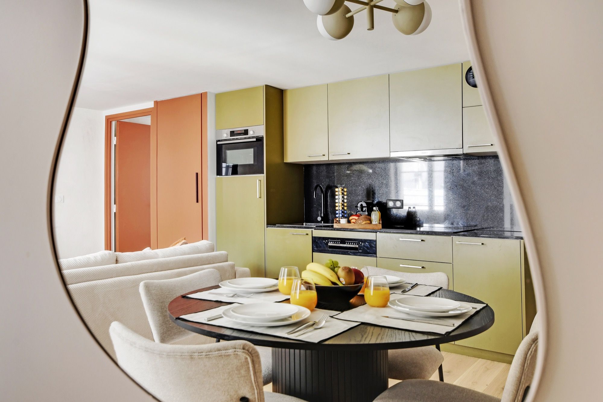 A modern kitchen with a dining area, featuring a round table set for four with plates, cutlery, and a fruit bowl.