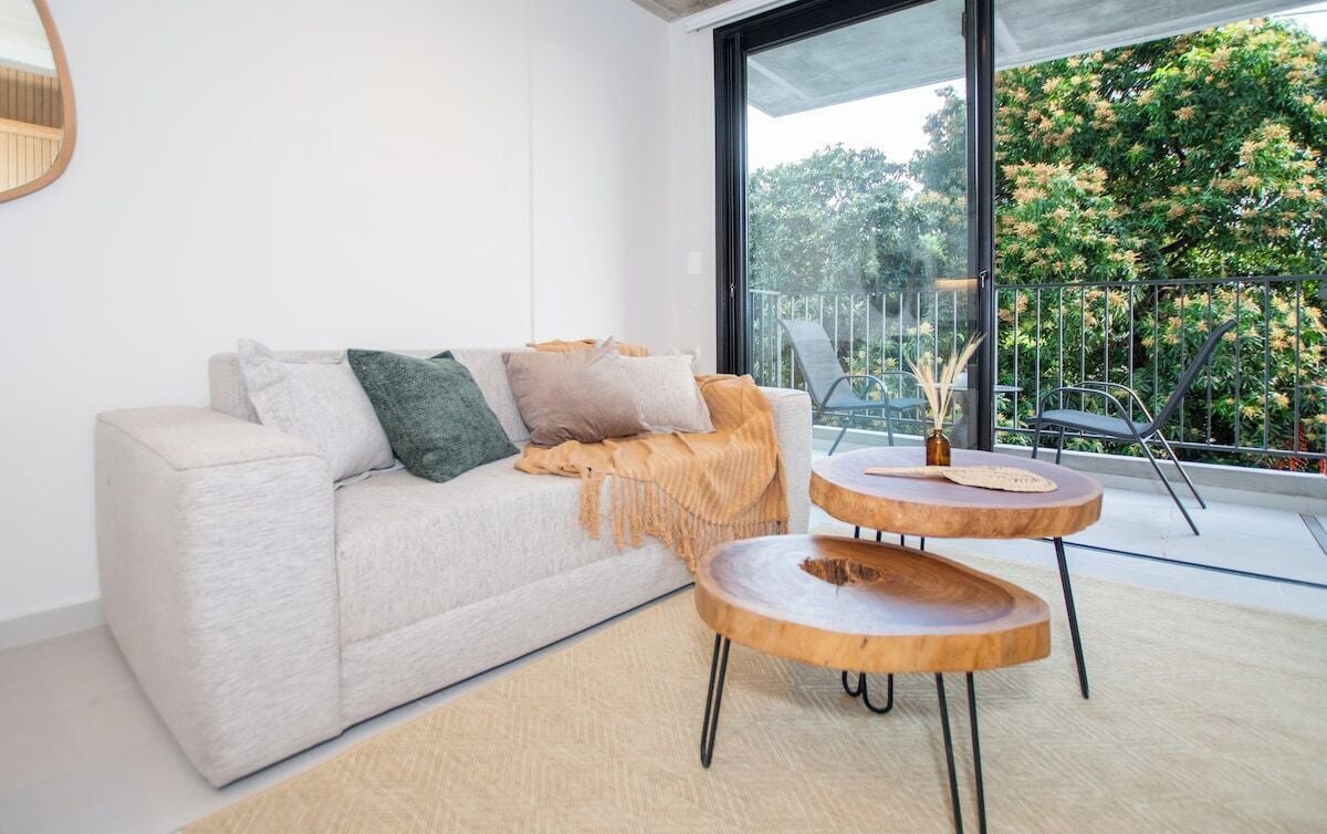 Un salon moderne avec un canapé, deux petites tables d’appoint en bois et une grande fenêtre donnant sur un paysage verdoyant.