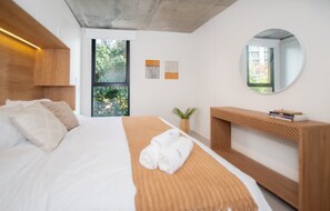 A modern bedroom with a large bed, a wooden console table, a round mirror, and a small potted plant. - Corporate Stays First del Sol - 106 (Asunción)