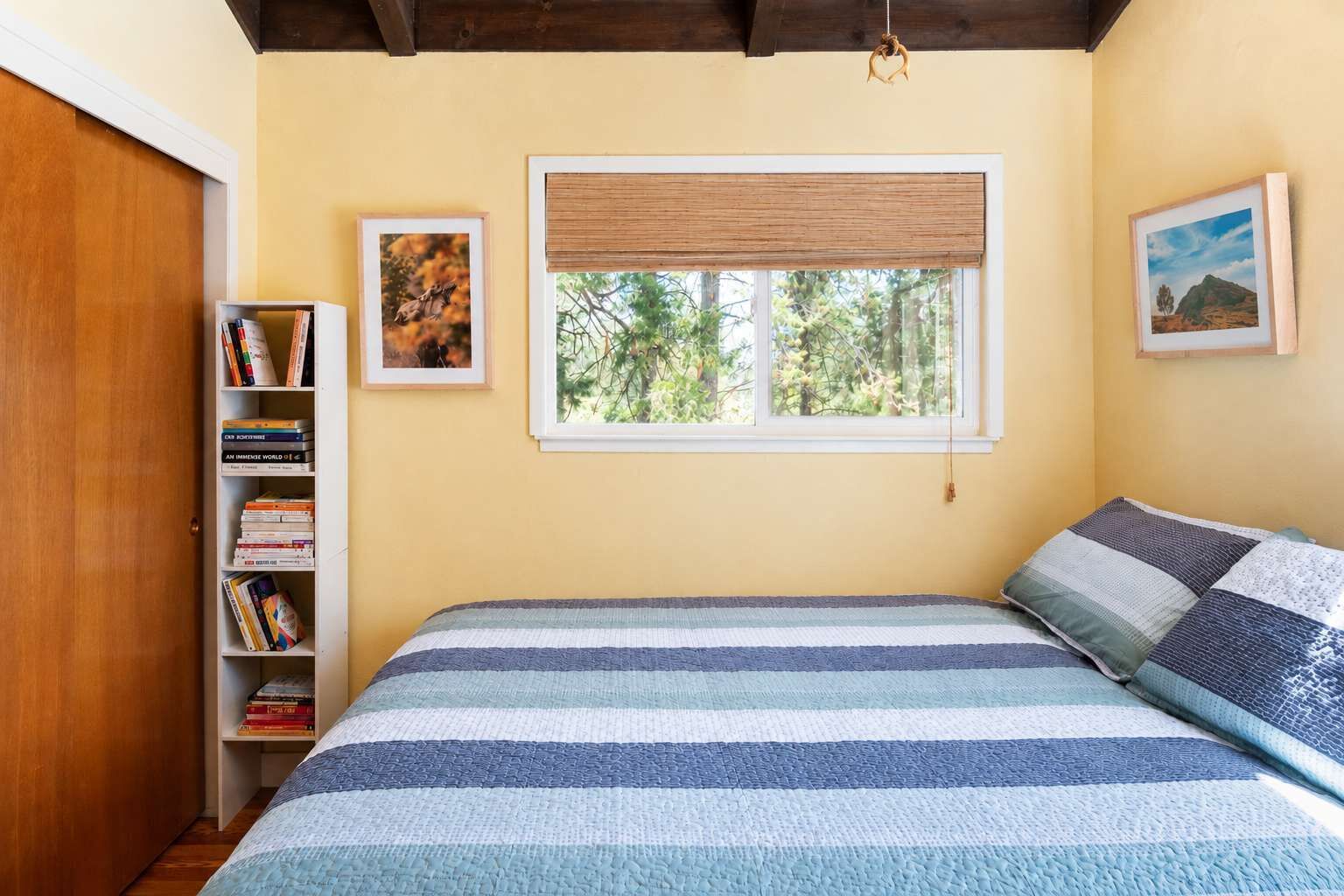 Een slaapkamer met een bed, een boekenkast, een raam met uitzicht op bomen en twee ingelijste foto's aan de muur.