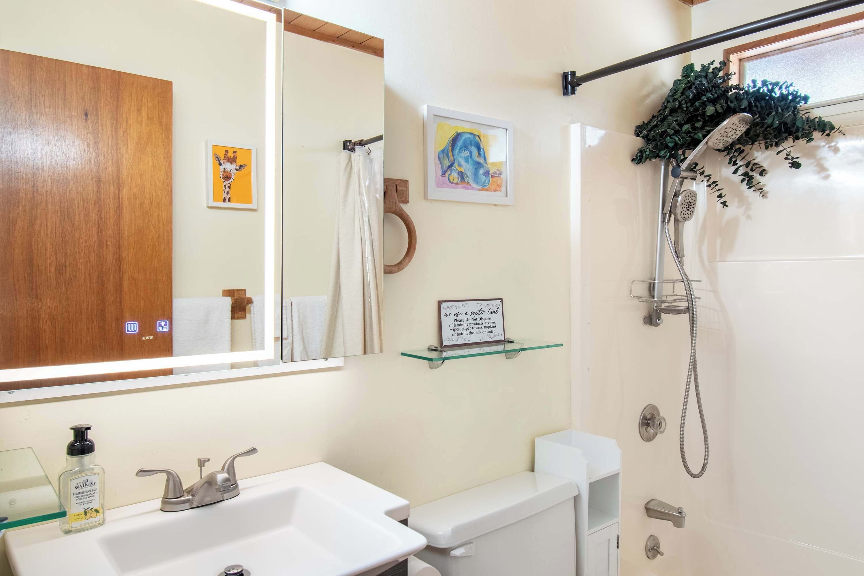 Een badkamer met een wit wastafelmeubel, een spiegel, een douche met een glazen plankje en een bordje over een septic tank.