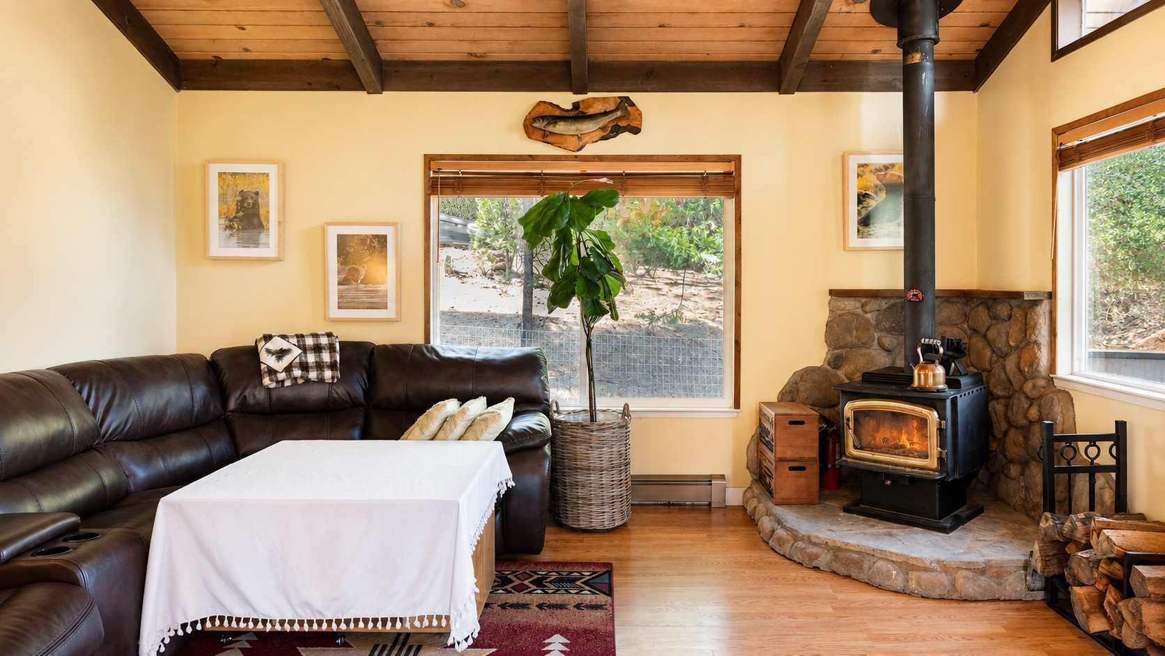 A cozy living room with a leather sofa, a table with a white tablecloth, a large plant, and a stone fireplace with a wood-burning stove.