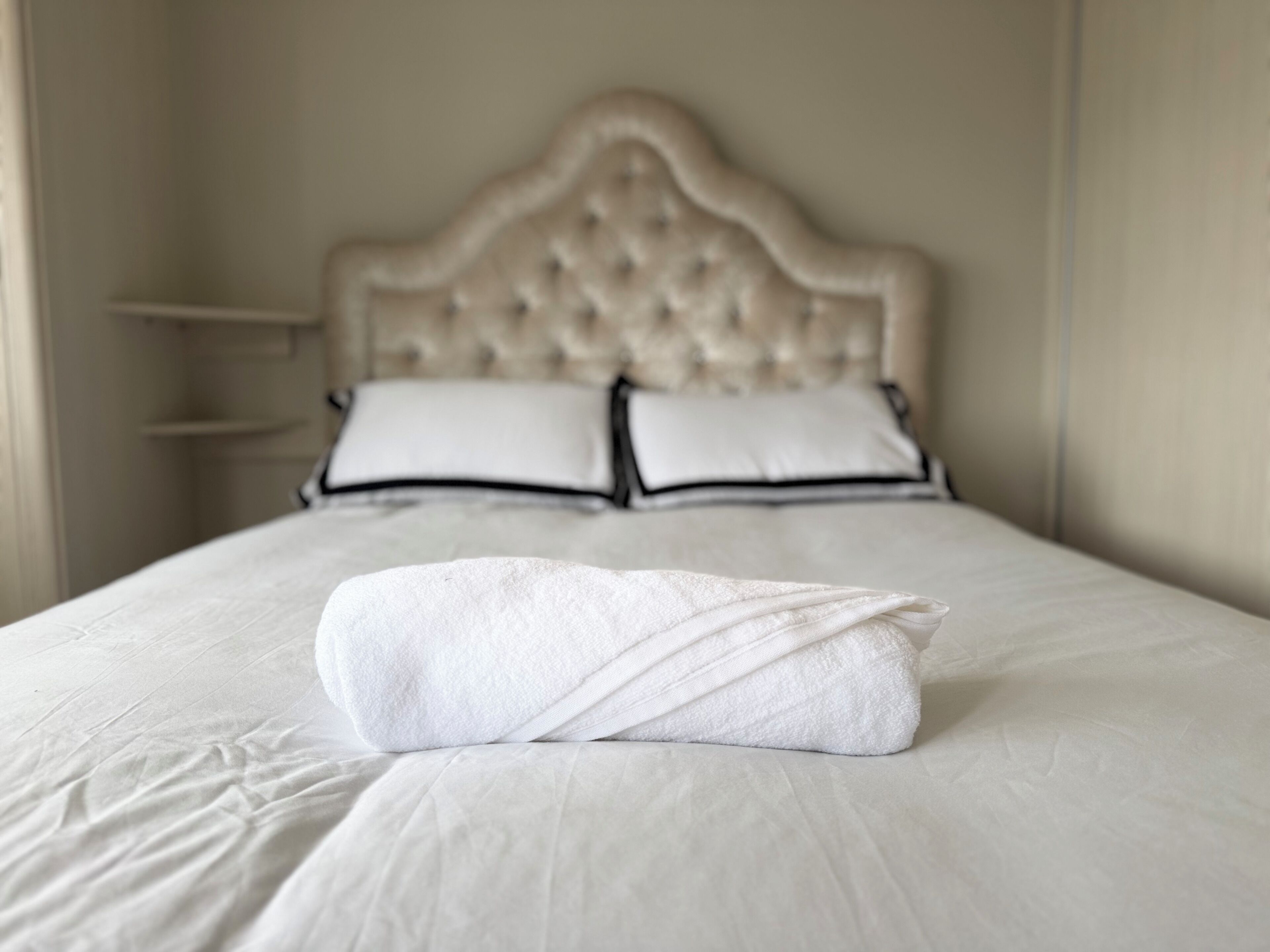 A neatly made bed with a folded white towel in the center, a headboard with a tufted design, and two pillows with black trim.
