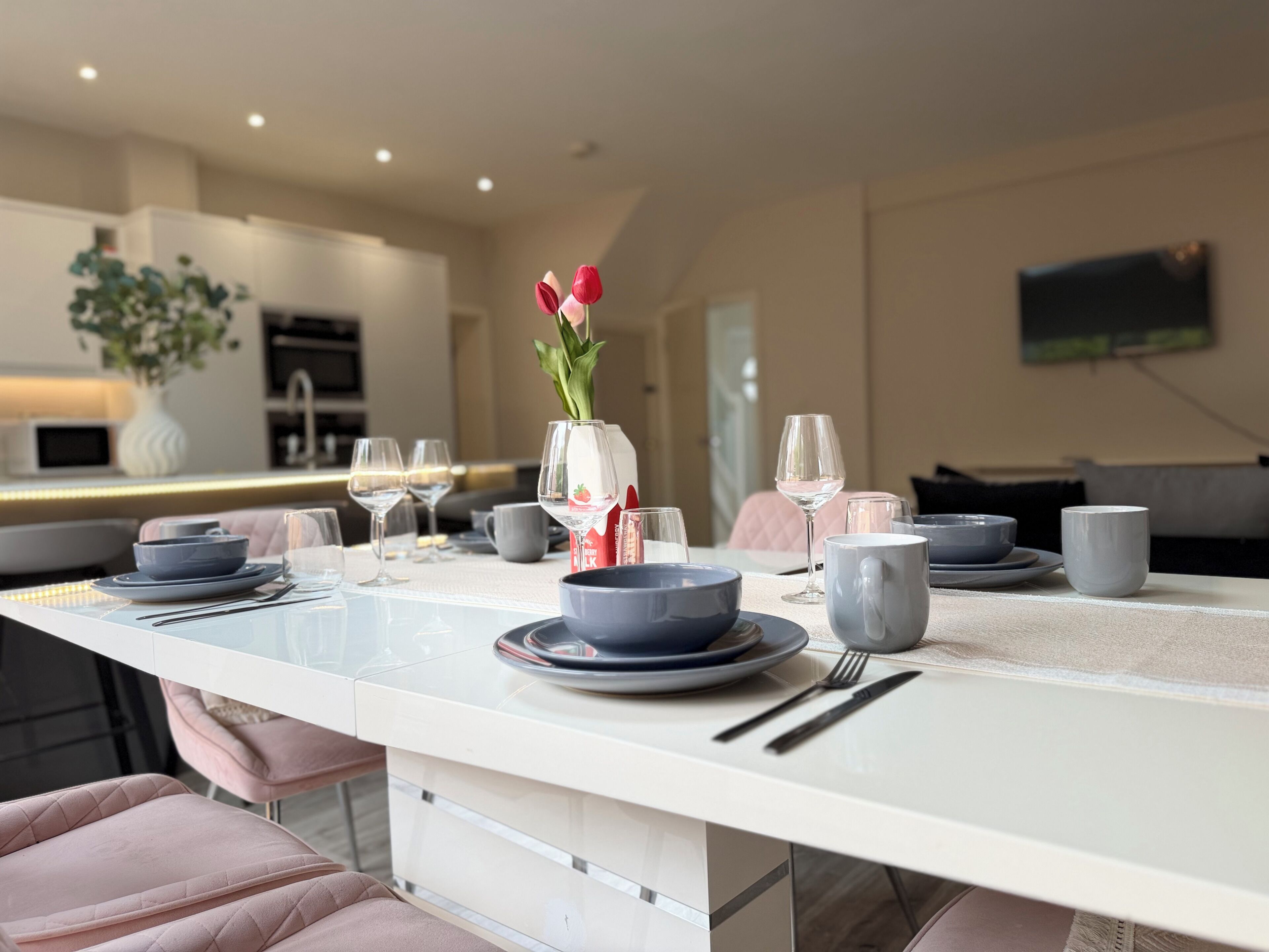 A modern dining area with a white table set for two, featuring a vase with flowers, wine glasses, and dinnerware.