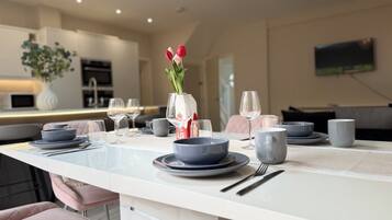 A modern dining area with a white table set for two, featuring a vase with flowers, wine glasses, and dinnerware.