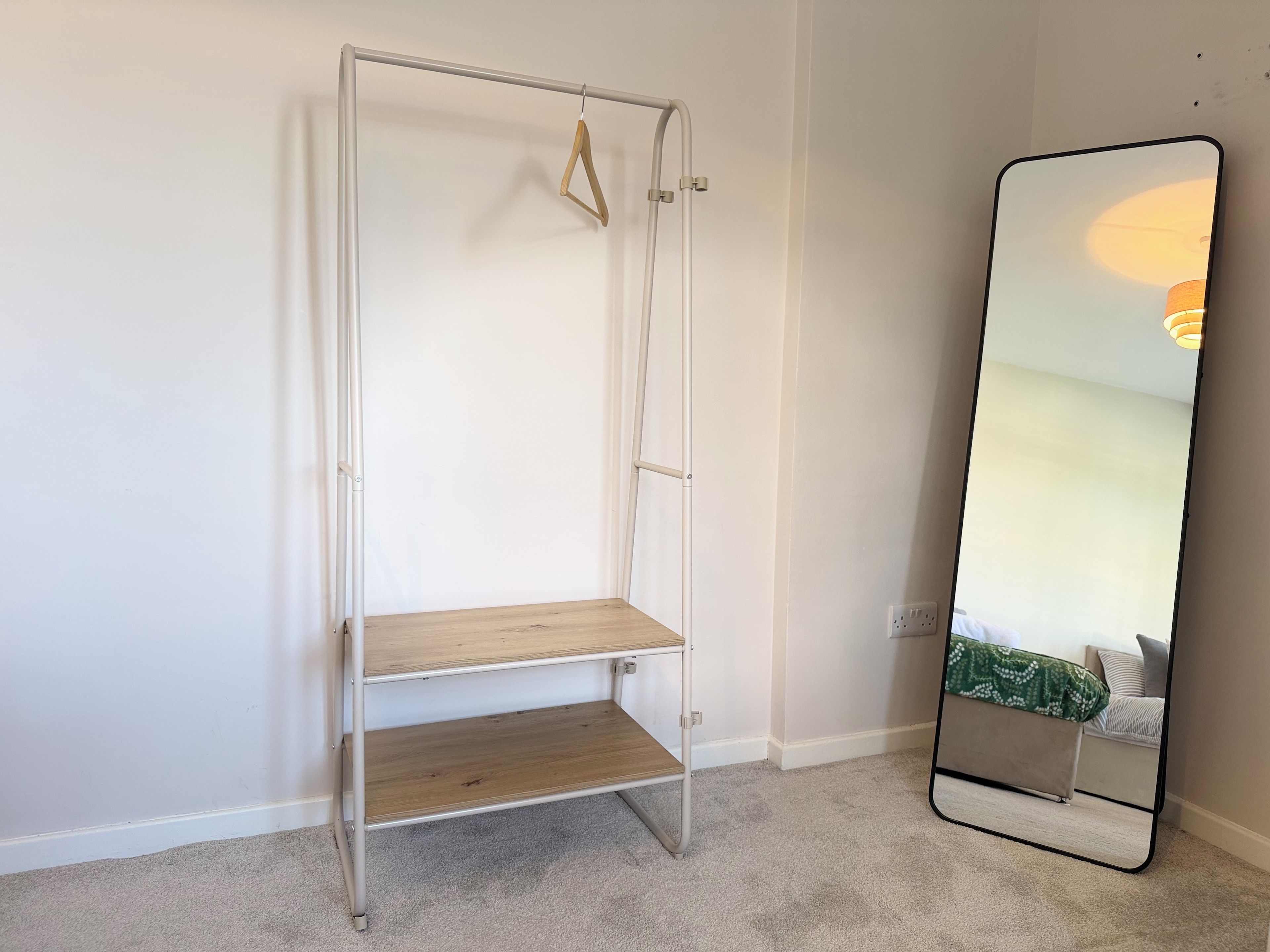 A room with a metal shelf unit against a wall, a carpeted floor, and a large mirror reflecting part of the room.