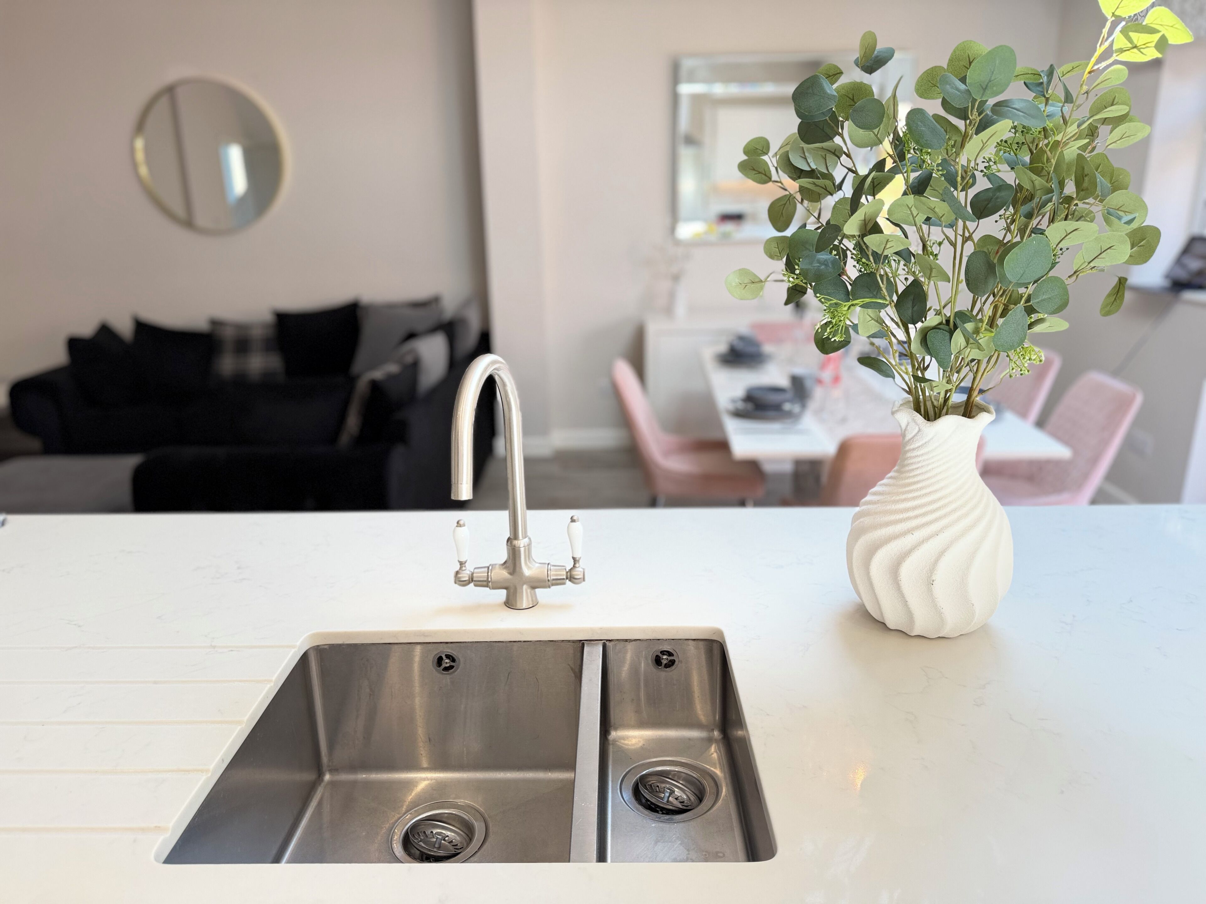 A kitchen with a double stainless steel sink, a white vase with green foliage, and a dining area in the background.
