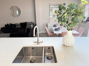 A kitchen with a double stainless steel sink, a white vase with green foliage, and a dining area in the background. - Spacious Family House-Private Hot Tub, Driveway & Easy Access to Beach & London (London)