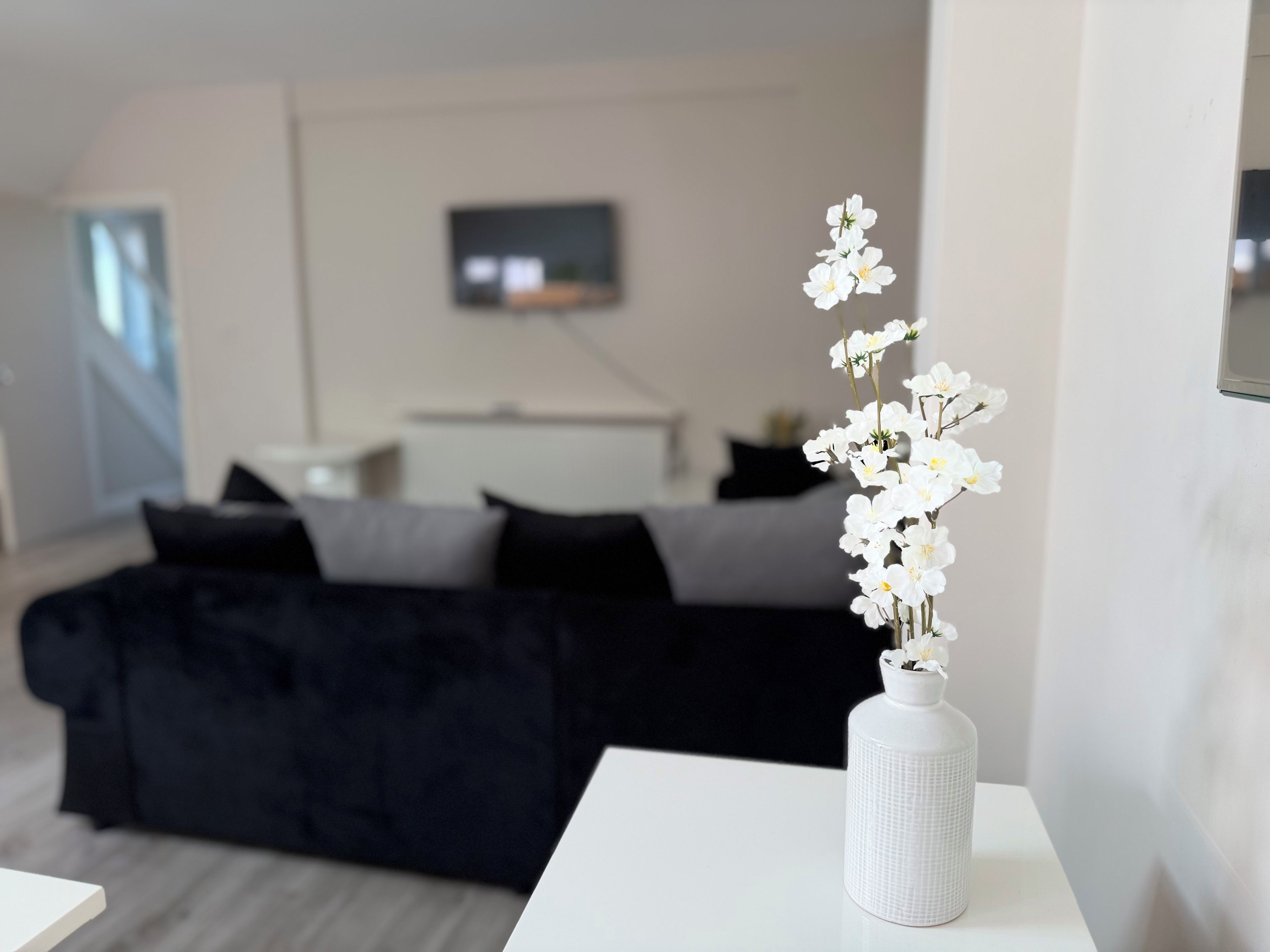 Quarto de hotel moderno com uma cama, uma TV de tela plana na parede e um vaso branco com flores brancas sobre uma mesa.