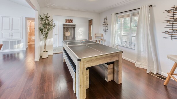 Um quarto espaçoso com mesa de ping pong, piso de madeira, uma planta em vaso e uma janela com persianas.