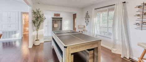 Amplia habitación con mesa de ping pong, piso de madera, planta en maceta y ventana con persianas.