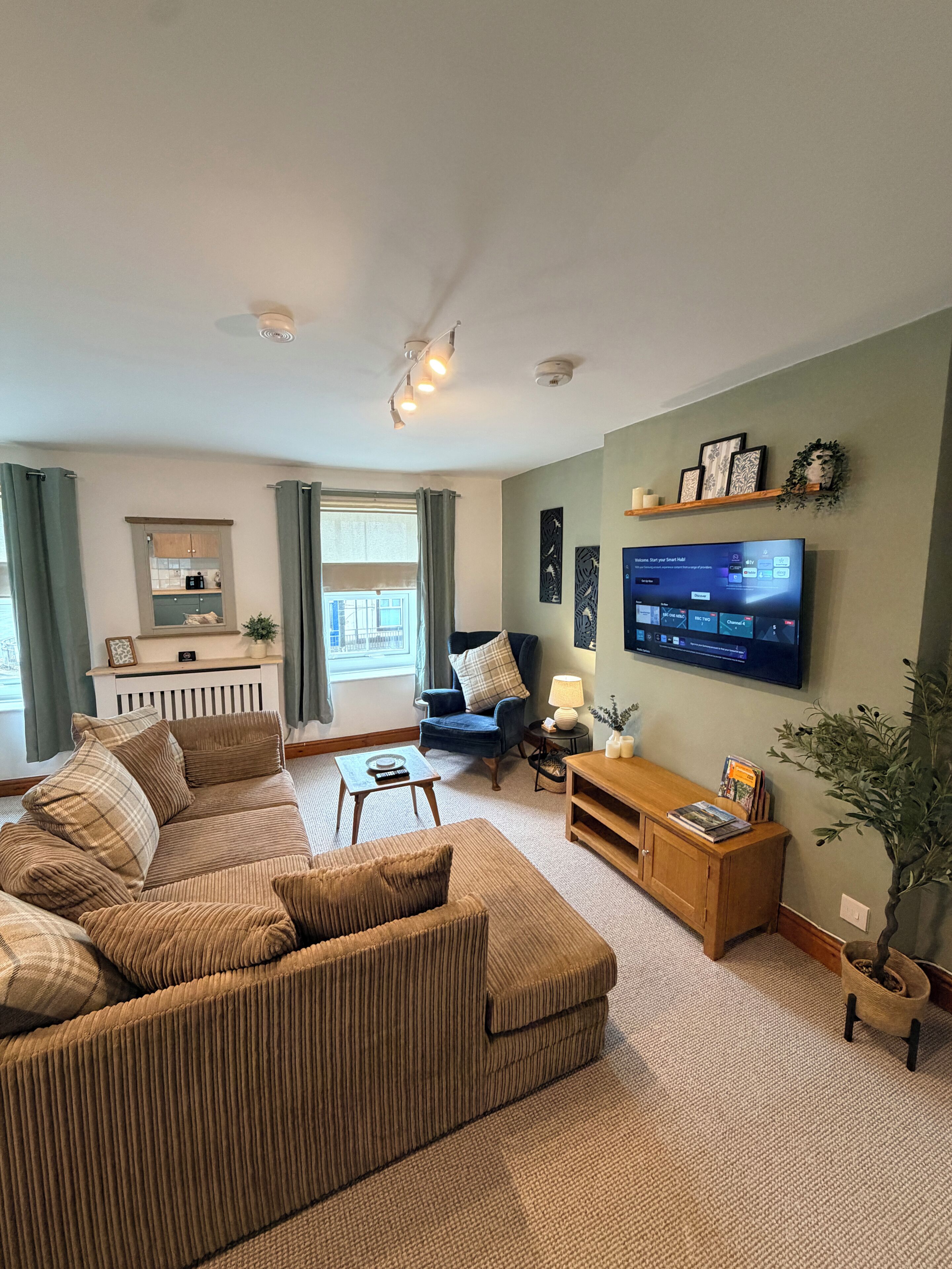 Een woonkamer met een bank, fauteuil, salontafel en een flatscreen televisie aan de muur.