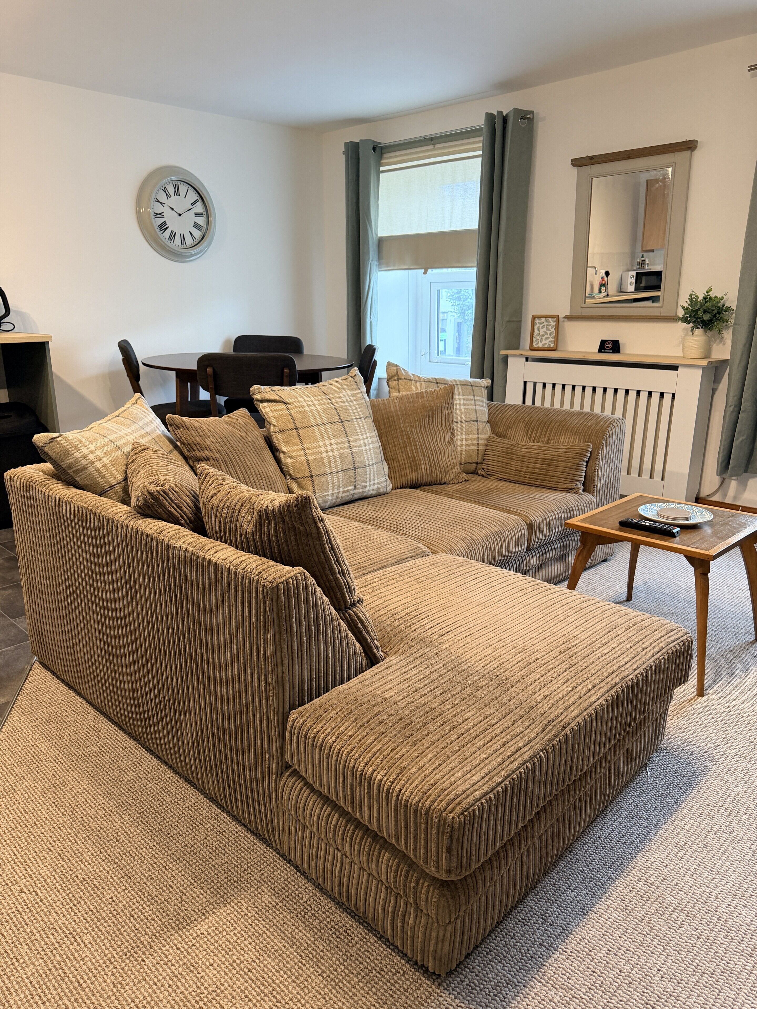 A living room with a sectional sofa, a dining table, chairs, a clock on the wall, a window with curtains, and a radiator.