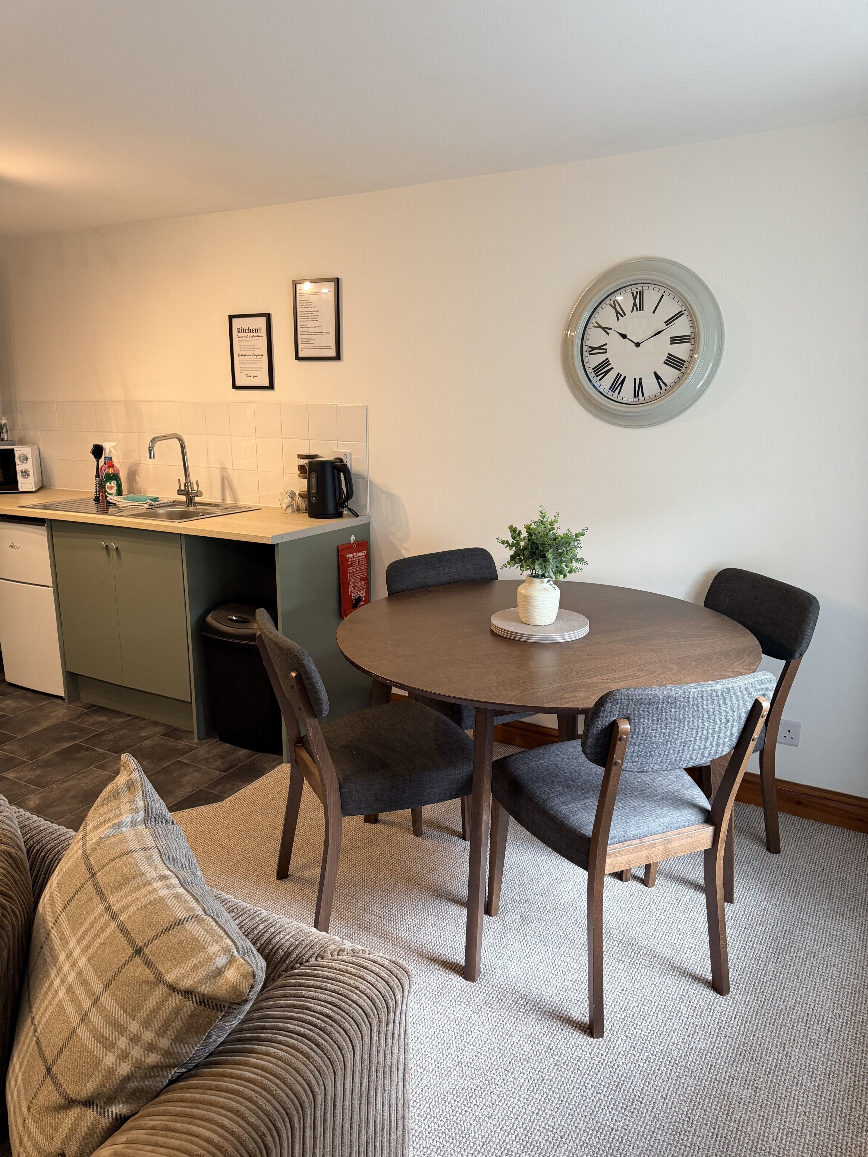 A modern kitchen with a sink, stove, and microwave. There is a dining table with chairs and a clock on the wall.