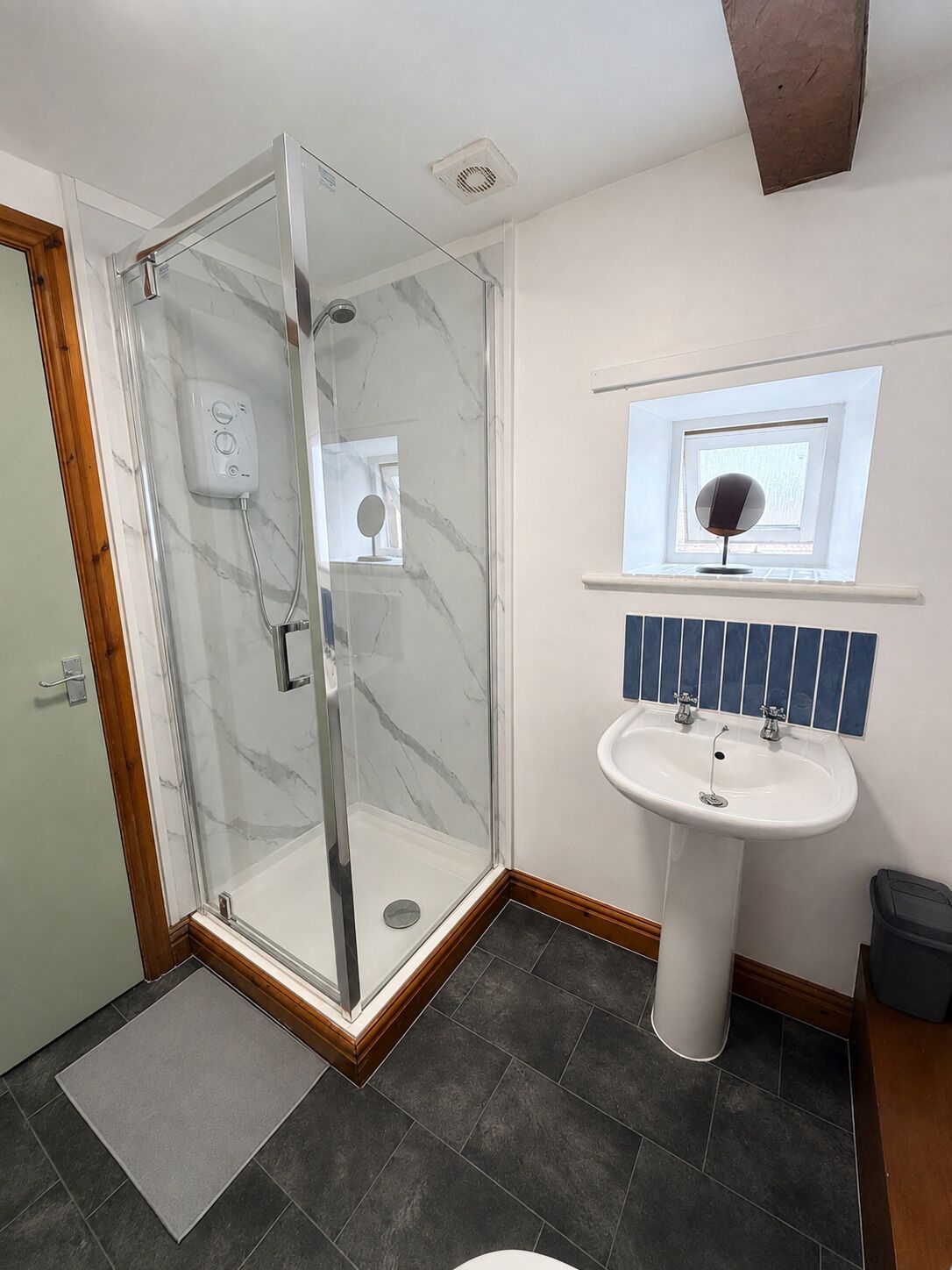 A modern bathroom with a glass-enclosed shower, a pedestal sink, and a small window above the sink.