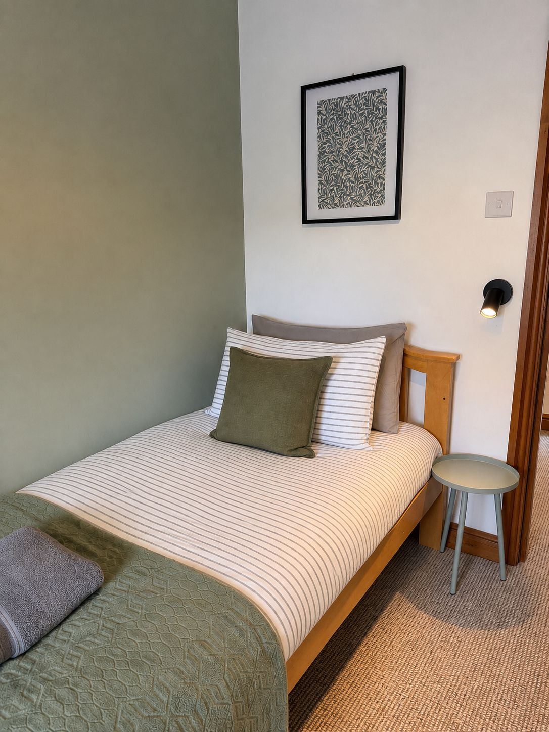 A single bed with a striped bedspread, a green throw blanket, and a pillow. There is a small round table beside the bed. A framed artwork hangs on the wall above the bed.