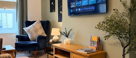 A living room with a television mounted on the wall, a blue armchair, a wooden coffee table, and a bookshelf.